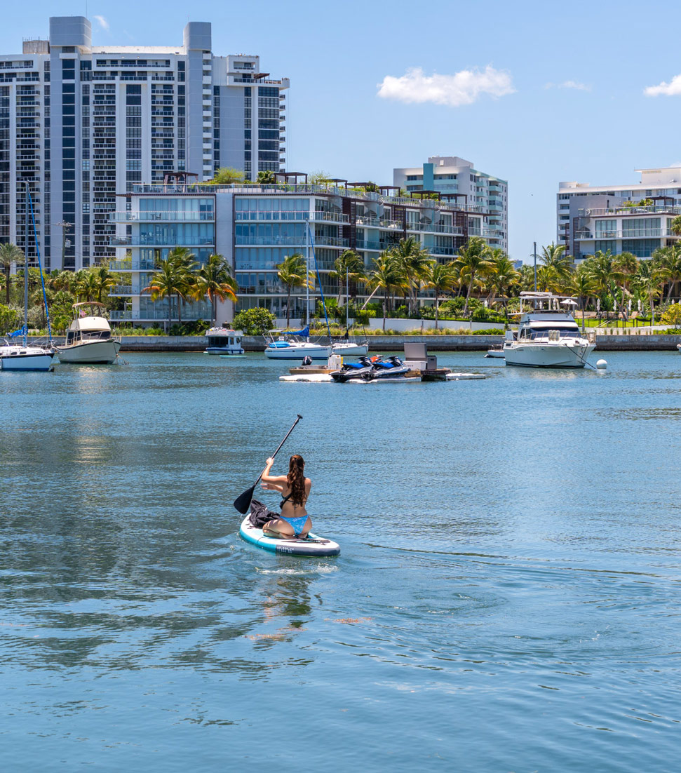 Woman paddleboarding on calm water with boats & buildings in the background
